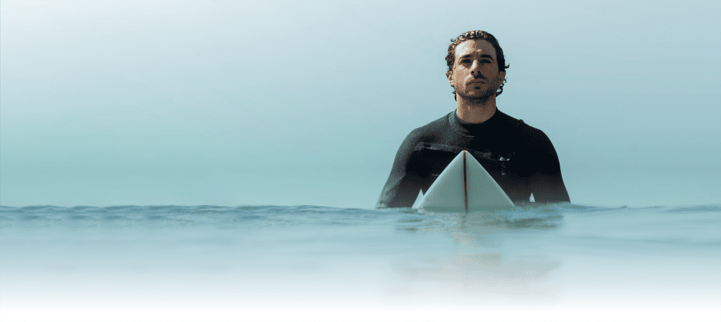 Surfista sendo na prancha dentro do mar. Indicando que está aguardando uma boa onda para surfar. Ele veste roupa apropriada para surfe de neoprene. Dia de cáu azul.