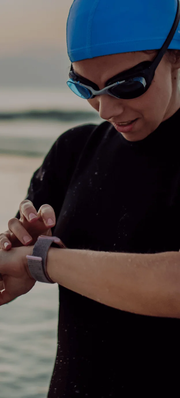 Mulher vestindo roupas de nadadora de neoprene, touca e óculos de natação. Olhando o relogio na praia.