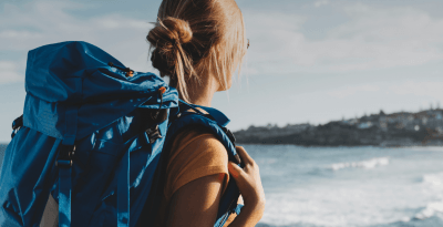 Mulher com mochila nas costas, indicando que está fazendo trilha, parada no costão olhando o mar.