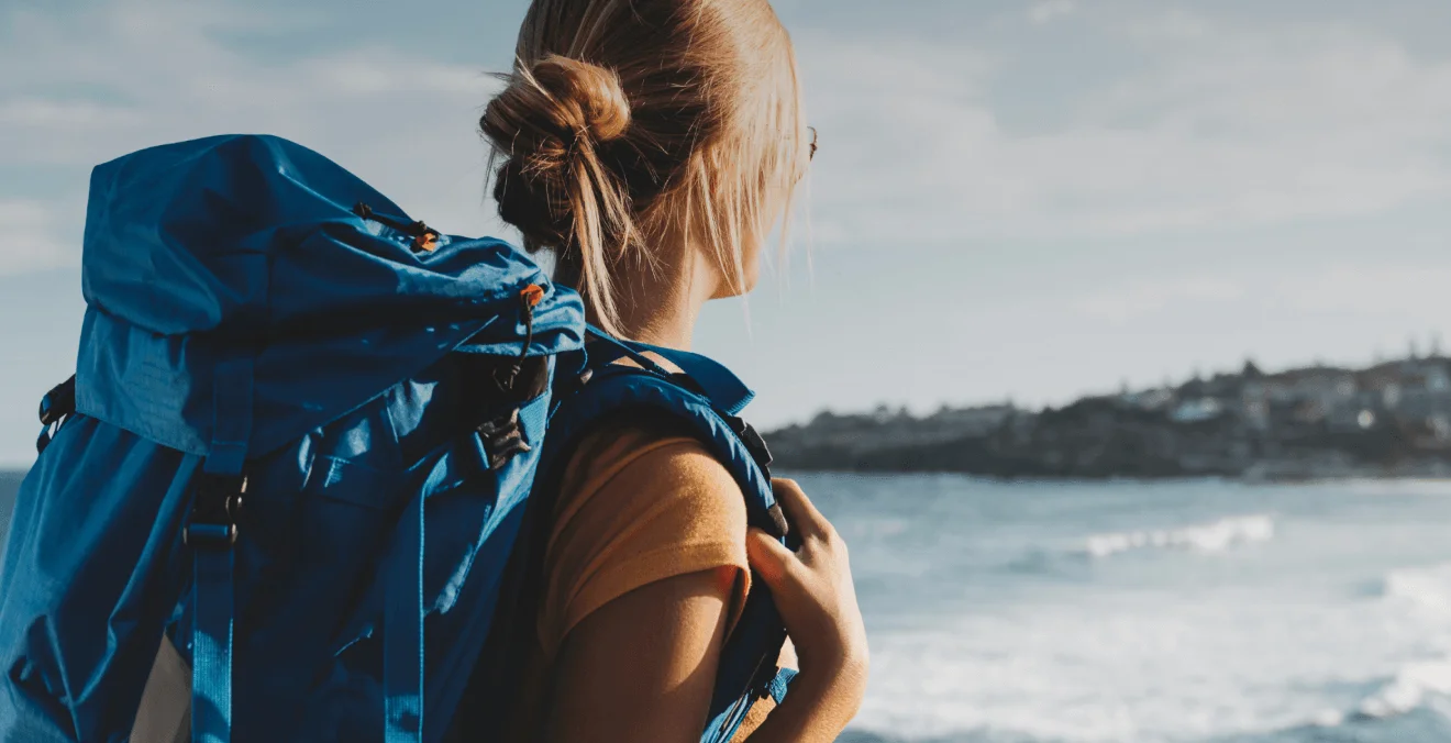 Mulher com mochila nas costas, indicando que está fazendo trilha, parada no costão olhando o mar.