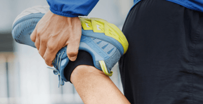 Pessoa alongando, puxando a perna atrás do gluteo. Usando moletom azul, bermuda preta e tenis azul