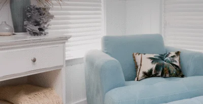 Quarto arejado e iluminado, paredes e móvel branco, poltrona azul piscina com almofada com imagem de palmeiras.