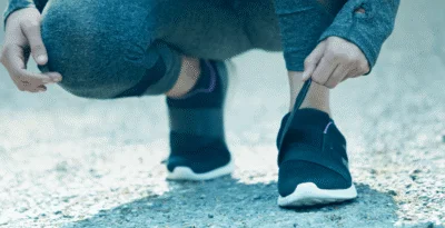 Pessoa agachada apertando o velcro do tenis. Vestindo roupas de caminhada e tenis preto.