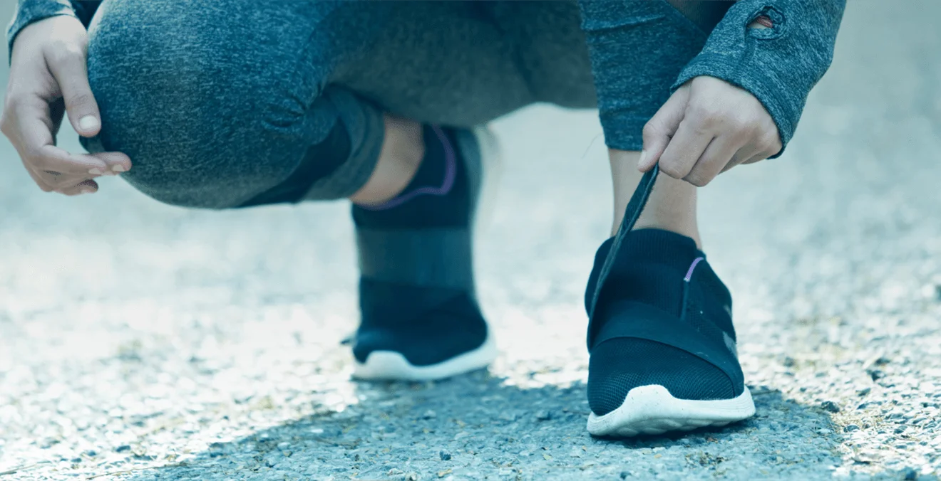 Pessoa agachada apertando o velcro do tenis. Vestindo roupas de caminhada e tenis preto.